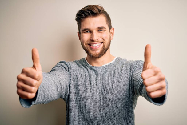ひげと青い目をした若いハンサムなブロンドの男は、手で積極的なジェスチャーを行うことを承認カジュアルセーターを着て、親指を上げて笑顔と成功のために幸せ。勝者のジェスチャー. - 写真・画像