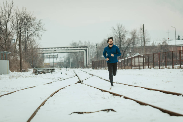 Een actieve jongeman rent en doet oefeningen over de oude spoorweg tijdens de wintertraining buiten.  - Foto, afbeelding