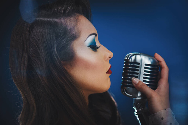 A beautiful woman singer with stylish retro hair and makeup is holding a vintage microphone with stand ready to sing. - Foto, afbeelding