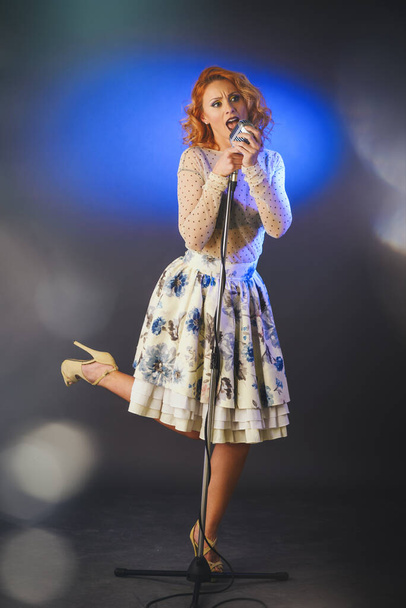 Красивая блондинка-певица со стильными ретро-волосами и макияжем держит винтажный микрофон с подставкой и поет
. - Фото, изображение
