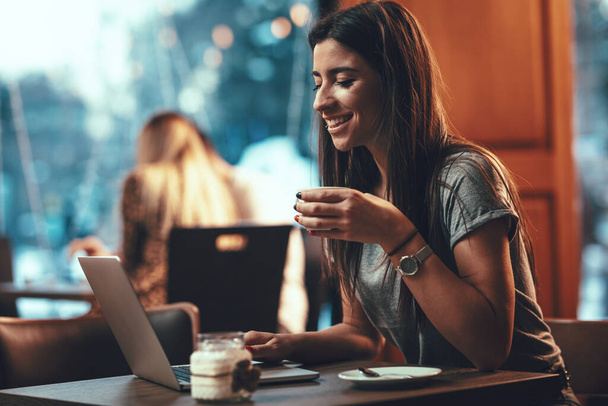 Houkutteleva nainen on onnellinen, kun lukee hyviä uutisia liikekumppaneilta kannettavalla tietokoneella, istuu ja työskentelee kahvilassa
. - Valokuva, kuva