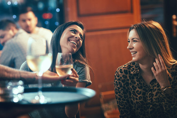 Sonrientes jóvenes queridas amigas están sentadas en el café, hablando y bebiendo vino
. - Foto, imagen