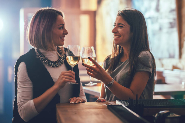 Lächelnde junge Freundinnen sitzen im Café, reden, stoßen an und trinken Wein.. - Foto, Bild