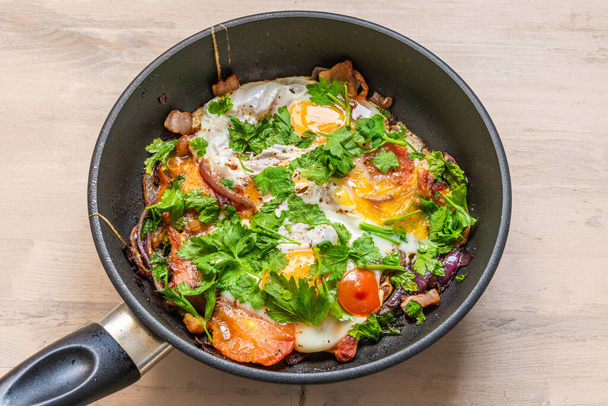 El desayuno fresco rústico - el huevo frito con el tocino, la cebolla y los tomates cherry en la cacerola
 - Foto, imagen