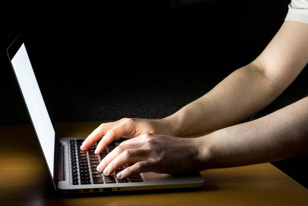 Человек работает на ноутбуке с белым экраном светится в темноте на деревянном столе видно со стороны с белой футболкой
 - Фото, изображение