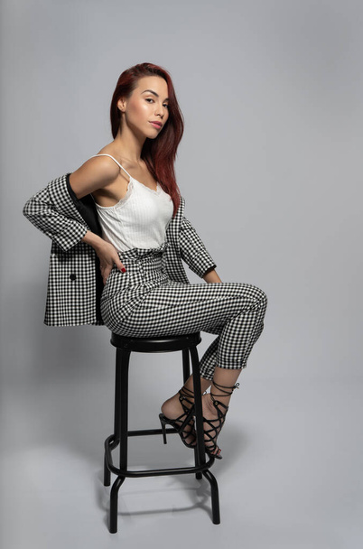 fashion shoot with red hair woman and black and white checkered suit and high heel shoe sitting on stool - Photo, Image