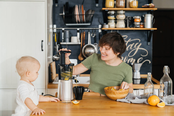 女性の母親と彼女の子供は朝のキッチンで一緒に新鮮なオレンジからおいしいジュースを作ります。家族のためのビタミンドリンク. - 写真・画像