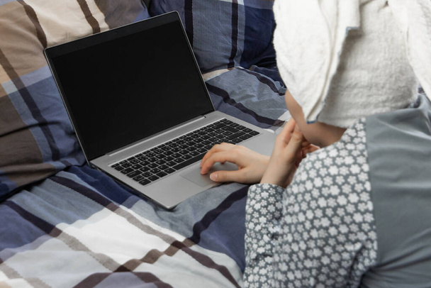 Залишайтеся вдома під час пандемії коронавірусу. Крупним планом жінка з загорнутим рушником на голові, використовуючи ноутбук у ліжку з блакитною постільною білизною
 - Фото, зображення