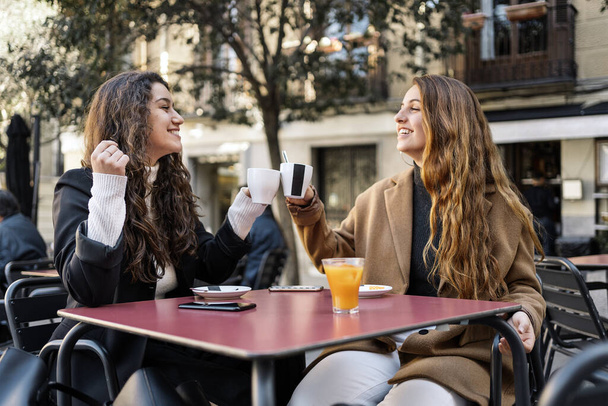 Zdjęcie dwóch młodych, kaukaskich kobiet rozmawiających siedząc w kawiarni. Uśmiechają się. Noszą eleganckie, luźne ubrania. Piją kawę.. - Zdjęcie, obraz