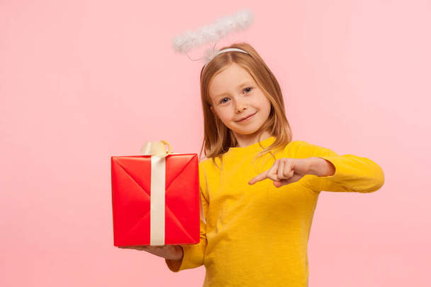 Portrait of fascinating kind little ginger girl with freckles and angelic halo pointing at gift box, congratulating on holiday, giving birthday present. indoor studio shot isolated on pink background - Photo, Image