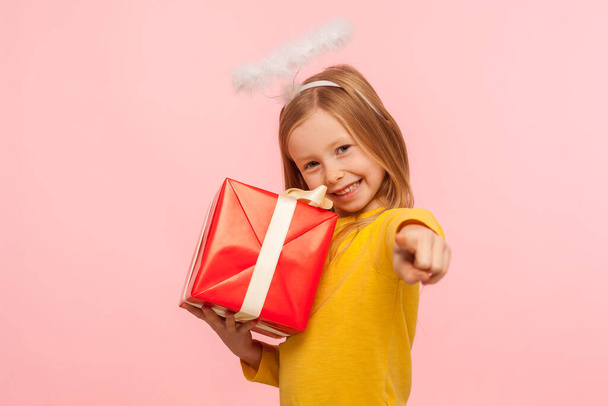 Hey, this present for you! Portrait of happy little ginger girl with angelic halo embracing wrapped box and pointing to camera, choosing lucky winner to give bonus, gift. indoor studio shot, isolated - Photo, image