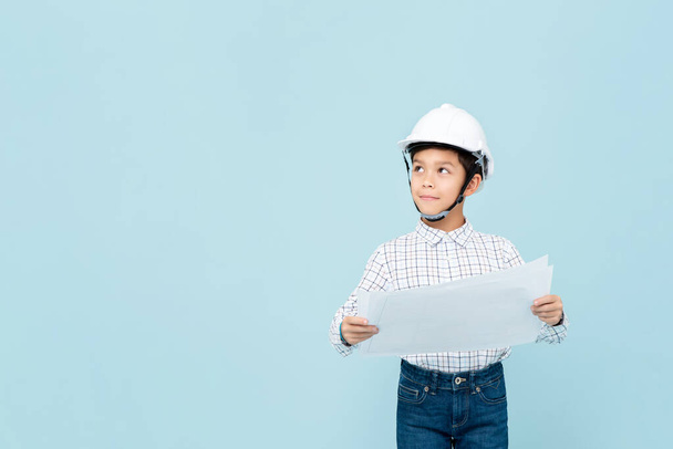 Smiling young asian boy dress like engineer with hardhat holding blueprint thinking and looking upward in light blue isolated studio backgorund - Photo, Image