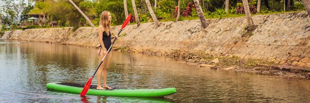 SUP青い水の上にパドルボード上の幸せな立って湖の上にパドルボードの女性のパドルボードを立ちます。パドルボード上の若い女性のアクションショットバナー,ロングフォーマット - 写真・画像