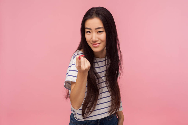 Come here! Portrait of attractive girl with brunette hair in striped t-shirt gesturing come to me, beckoning with finger, inviting for confidential talk. indoor studio shot isolated on pink background - Photo, image