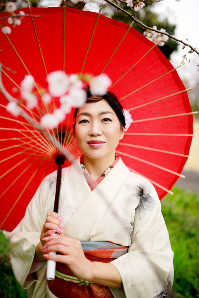 Eine Japanerin im weißen Kimono hält einen roten Regenschirm in der Hand und genießt den Blick auf die Kirschblüten in Tokio, Japan. - Foto, Bild