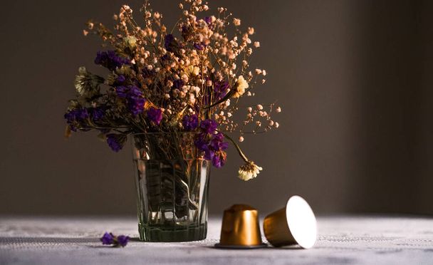 Две капсулы кофе Nespresso на столе перед фиолетовыми и белыми цветами в стеклянной вазе на белом текстильном фоне на темном фоне
 - Фото, изображение