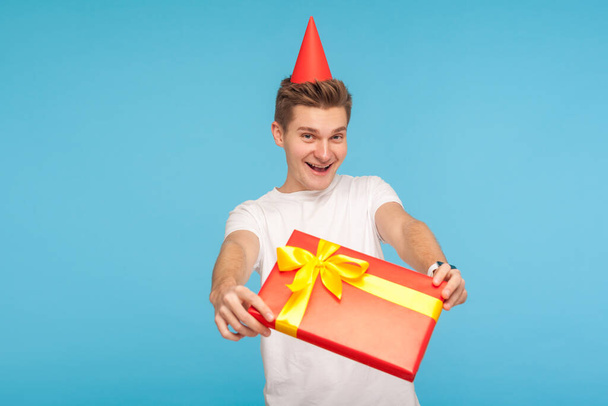 Surprise! Present for you! Funny glad generous man in white t-shirt and party cone on head giving gift box and smiling with childish carefree expression. indoor studio shot isolated on blue background - Foto, Imagem