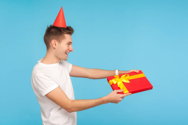 Take this present! Side view of funny cheerful generous man in t-shirt and party cone on head giving gift box and smiling sincerely, congratulating on holiday. indoor studio shot blue background - Фото, изображение