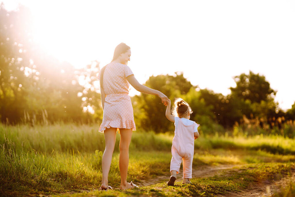 母と娘は日没の夏の公園で一緒に遊んでいます。日当たりの良いフィールドで楽しんでいる小さな子供と幸せなお母さん。キスと抱擁。幸せな家庭の概念.  - 写真・画像