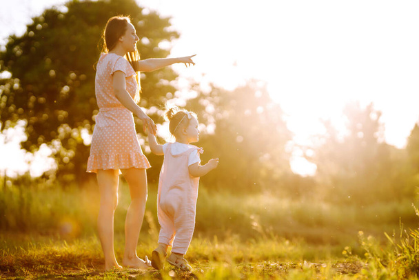 Matka i córeczka bawią się razem w letnim parku o zachodzie słońca. Szczęśliwa mama z małym dzieckiem bawiącym się na słonecznym polu. Całusy i uściski. Koncepcja szczęśliwej rodziny.  - Zdjęcie, obraz