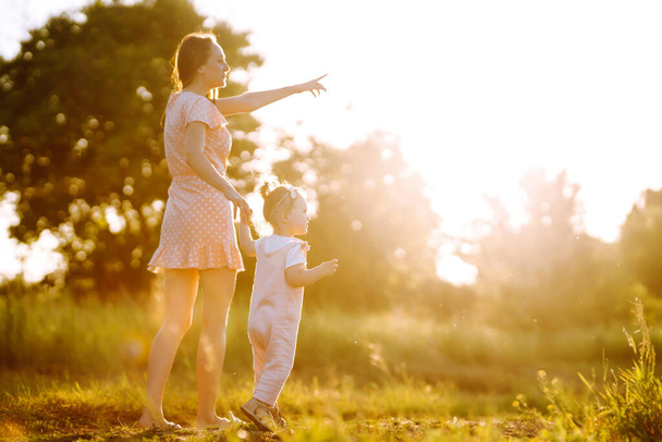 Mother and little daughter playing together in a summer park at sunset. Happy mom with small child  having fun on sunny field. Kisses and hugs. The concept of a happy family.  - 写真・画像