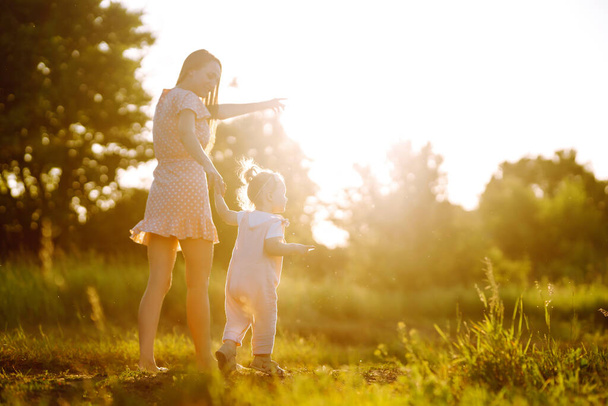 Μητέρα και μικρή κόρη παίζουν μαζί σε ένα καλοκαιρινό πάρκο το ηλιοβασίλεμα. Ευτυχισμένη μαμά με μικρό παιδί που διασκεδάζει στο ηλιόλουστο γήπεδο. Φιλιά και αγκαλιές. Η έννοια της ευτυχισμένης οικογένειας.  - Φωτογραφία, εικόνα