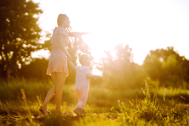 Mother and little daughter playing together in a summer park at sunset. Happy mom with small child  having fun on sunny field. Kisses and hugs. The concept of a happy family.  - Фото, зображення