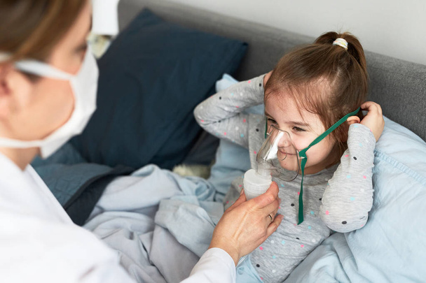 Doctor visiting little patient at home. Child having medical inhalation treatment with nebuliser. Girl with breathing mask on her face. Woman wearing uniform and face mask - Foto, afbeelding