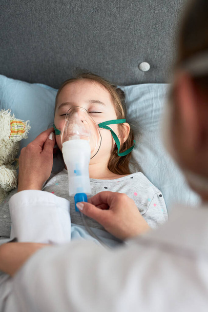 Doctor visiting little patient at home. Child having medical inhalation treatment with nebuliser. Girl with breathing mask on her face. Woman wearing uniform and face mask - Foto, immagini