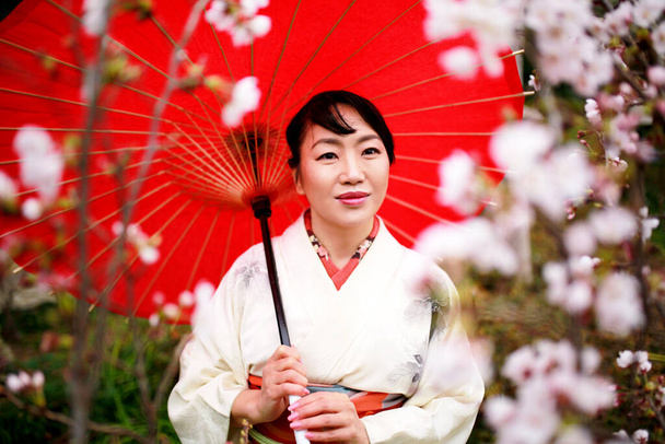 Японская женщина в белом кимоно держит красный зонтик, наслаждаясь видом цветущей сакуры в Токио, Япония
. - Фото, изображение