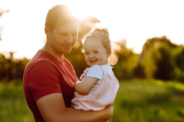 Ευτυχισμένος μπαμπάς και κοριτσάκι διασκεδάζουν στο πάρκο το ηλιοβασίλεμα. Ο μπαμπάς παίζει με ένα μικρό παιδί στο ηλιόλουστο γήπεδο. Φιλιά και αγκαλιές πατέρα και κόρης. Οικογενειακή έννοια και έννοια πατρότητας. - Φωτογραφία, εικόνα