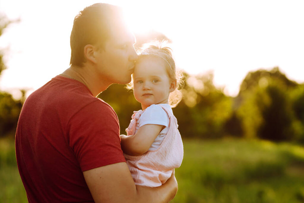 Happy dad and little baby girl  having fun in summer park at sunset. Dad plays with small child on sunny field. Kisses and hugs of father and daughter. Family  and fatherhood concept. - Foto, imagen