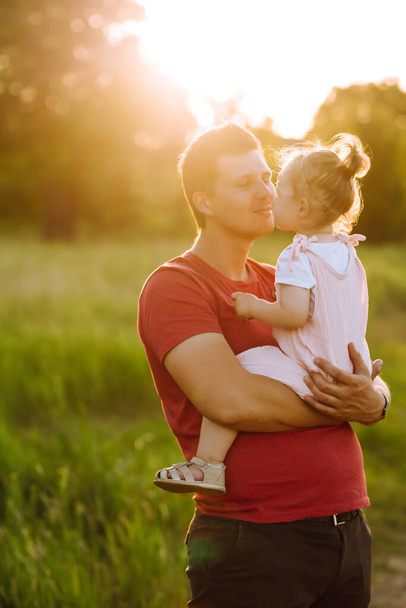 Happy dad and little baby girl  having fun in summer park at sunset. Dad plays with small child on sunny field. Kisses and hugs of father and daughter. Family  and fatherhood concept. - Photo, image