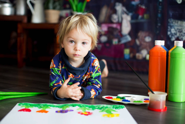 Γλυκό μικρό ξανθό παιδί, αγόρι, ζωγραφίζει με χρώματα, μικρά πουλάκια περπατούν γύρω του, κάνοντας αστεία αποτυπώματα στο χαρτί του. - Φωτογραφία, εικόνα