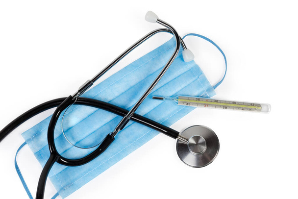 Стетоскоп и классический ртутный клинический термометр лежат на одноразовой медицинской маске, вид сверху
 - Фото, изображение