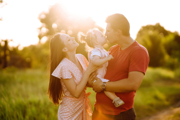 Happy young family walking in the park at sunset. Mom, dad and little daughter having fun in summer park. The concept of a happy family. Parents hold the baby's hands. Kisses and hugs. - Фото, зображення
