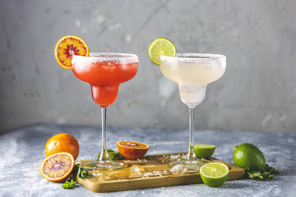 Frozen lime margarita and blood orange margarita cocktail mix in salt rimmed glasses garnished with slices of lime and orange. Focus on the citrus slice. Shallow depth of the field - Φωτογραφία, εικόνα