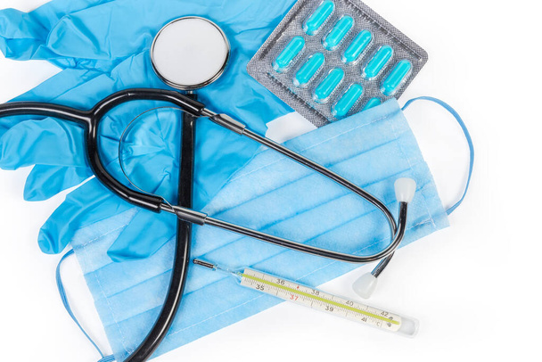 Stethoscoop en klassieke kwik klinische thermometer liggen op het wegwerp medisch masker en nitril medische handschoenen, blisterverpakking van pillen op een witte achtergrond, bovenaanzicht - Foto, afbeelding