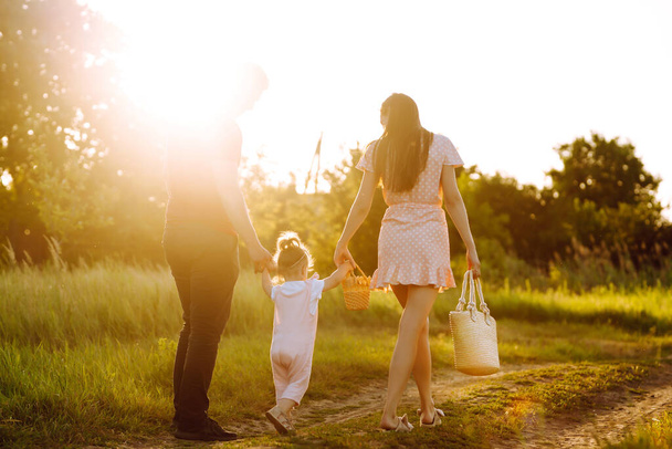 Szczęśliwa młoda rodzina spacerująca po parku o zachodzie słońca. Mama, tata i córeczka bawią się w letnim parku. Koncepcja szczęśliwej rodziny. Rodzice trzymają dziecko za ręce. Pocałunki i uściski. - Zdjęcie, obraz