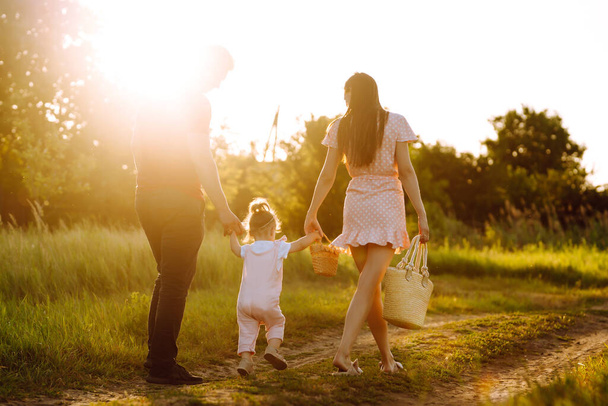 Ευτυχισμένη οικογένεια που περπατά στο πάρκο το ηλιοβασίλεμα. Η μαμά, ο μπαμπάς και η μικρή κόρη διασκεδάζουν στο πάρκο. Η έννοια της ευτυχισμένης οικογένειας. Οι γονείς κρατούν τα χέρια του μωρού. Φιλιά και αγκαλιές. - Φωτογραφία, εικόνα