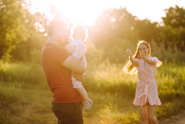 Šťastná mladá rodina procházející se v parku při západu slunce. Máma, táta a dcerka se baví v letním parku. Koncept šťastné rodiny. Rodiče drží dítě za ruce. Pusy a objetí. - Fotografie, Obrázek