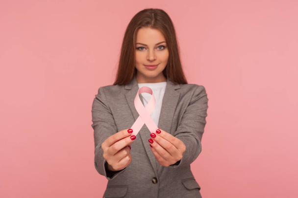 Female health, medical insurance. Portrait of young positive businesswoman in elegant suit jacket holding pink ribbon, warning of breast cancer risk and society awareness. studio shot, pink background - Foto, Bild