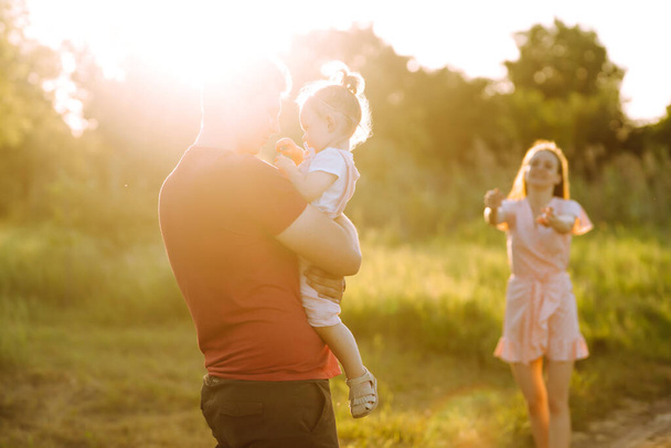 Boldog fiatal család sétál a parkban naplementekor. Anya, apa és a kislánya jól érzik magukat a nyári parkban. A boldog család fogalma. A szülők fogják a baba kezét. Csók és ölelés. - Fotó, kép