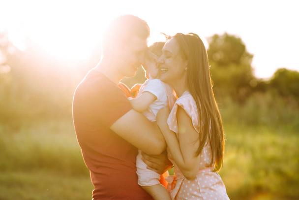 Happy young family walking in the park at sunset. Mom, dad and little daughter having fun in summer park. The concept of a happy family. Parents hold the baby's hands. Kisses and hugs. - Φωτογραφία, εικόνα