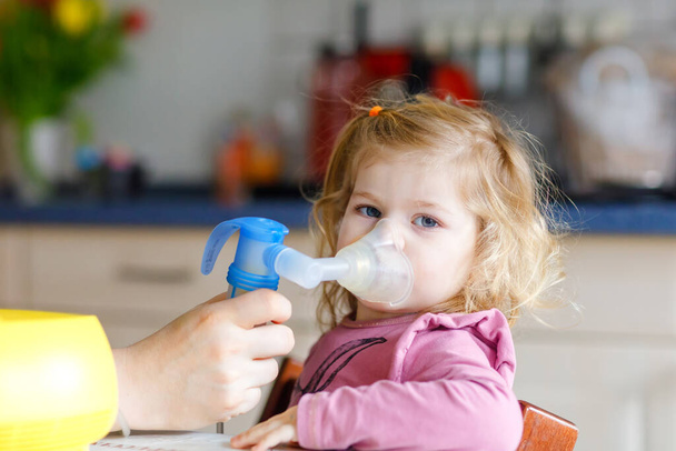 Küçük kız evde nebulizörle nefes alıp veriyor. Baba ya da anne yardım ediyor ve cihazı tutuyor. Çocuk grip, öksürük ve bronşit olmuş. Astım spreyi soluması buhar tutması konsepti - Fotoğraf, Görsel