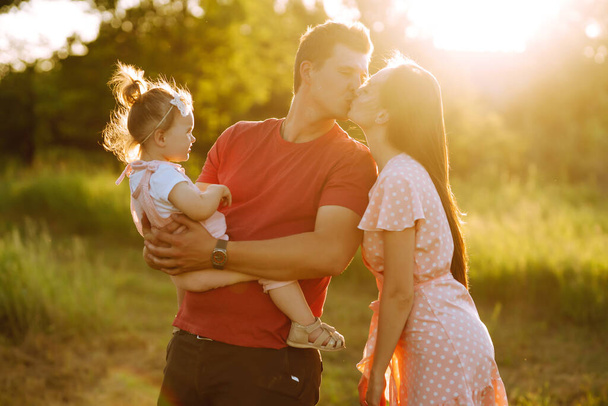 Happy young family walking in the park at sunset. Mom, dad and little daughter having fun in summer park. The concept of a happy family. Parents hold the baby's hands. Kisses and hugs. - 写真・画像