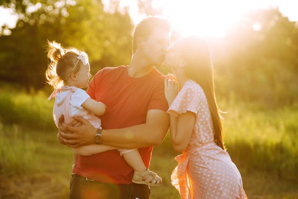 Happy young family walking in the park at sunset. Mom, dad and little daughter having fun in summer park. The concept of a happy family. Parents hold the baby's hands. Kisses and hugs. - Fotografie, Obrázek