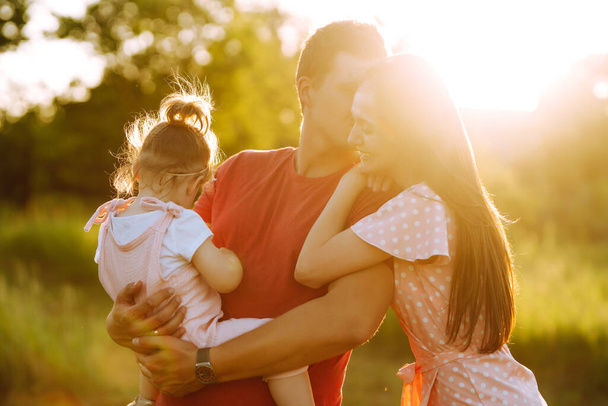 Happy young family walking in the park at sunset. Mom, dad and little daughter having fun in summer park. The concept of a happy family. Parents hold the baby's hands. Kisses and hugs. - Foto, Bild