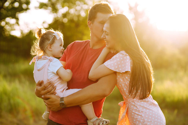 Happy young family walking in the park at sunset. Mom, dad and little daughter having fun in summer park. The concept of a happy family. Parents hold the baby's hands. Kisses and hugs. - Foto, Imagen