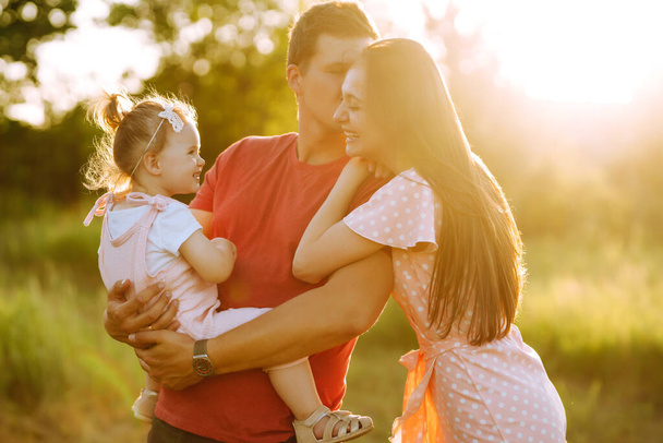 Happy young family walking in the park at sunset. Mom, dad and little daughter having fun in summer park. The concept of a happy family. Parents hold the baby's hands. Kisses and hugs. - Foto, afbeelding
